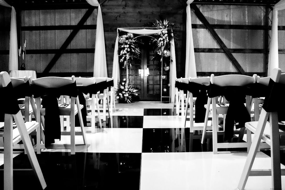 A wedding ceremony set up at an Oklahoma event venue — a floral arch at the end of a black-and-white checkerboard aisle lined with white chairs.